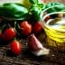 Cherry tomatoes, garlic, fresh basil, and a small bowl of olive oil on a wooden surface. - Olive Oil Times