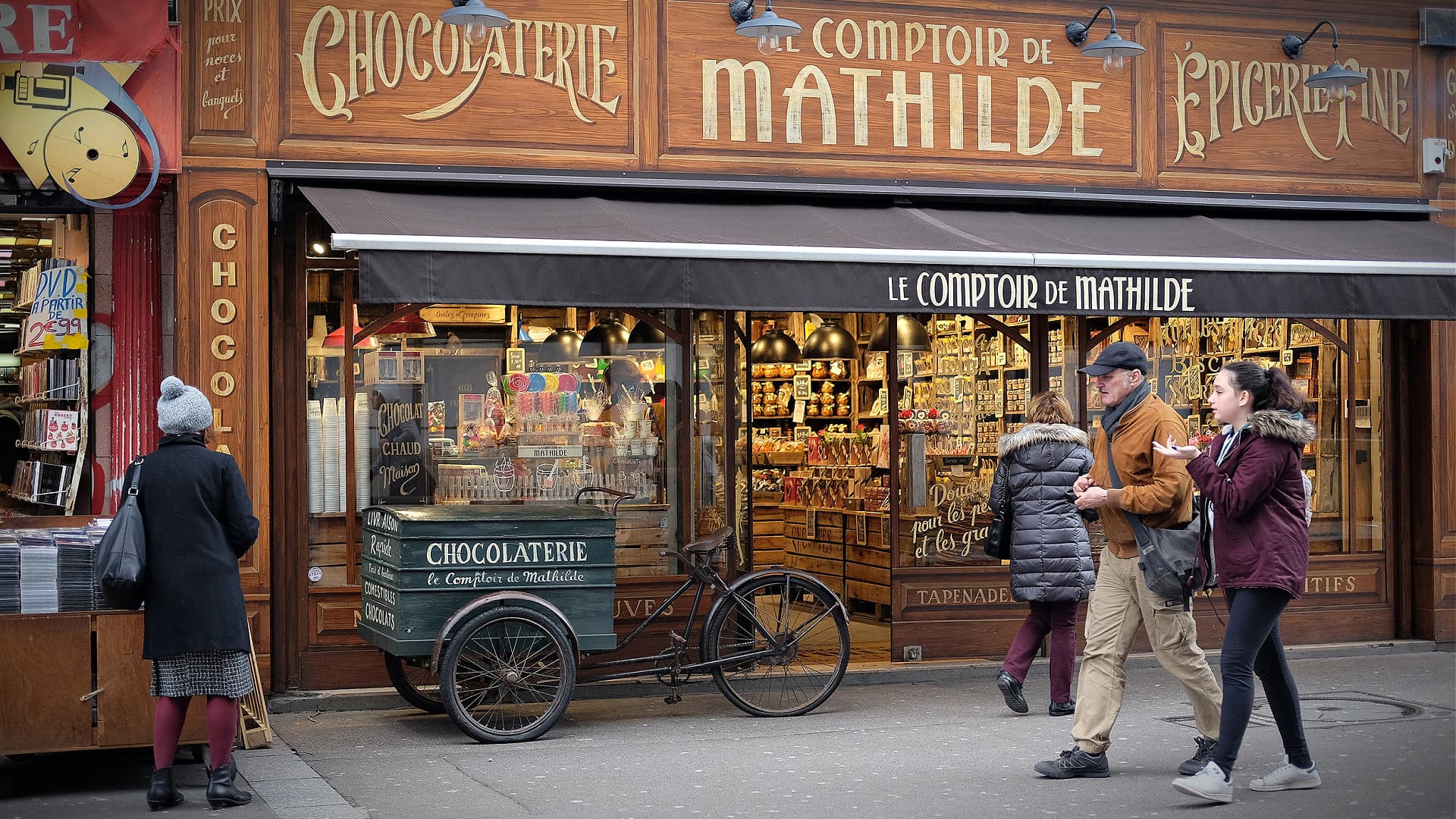 Shopfront of Le Comptoir de Mathilde featuring a chocolatier display and a bicycle cart. - Olive Oil Times
