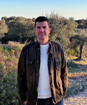 A man wearing a dark jacket and white sweater standing in an olive grove with trees in the background. - Olive Oil Times
