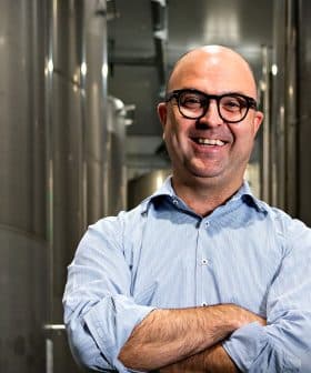 A man with glasses smiling and standing with arms crossed in a production facility surrounded by large metal tanks. - Olive Oil Times