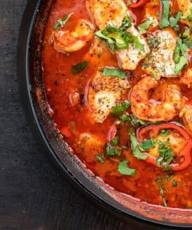A black dish containing chicken pieces, red peppers, and herbs in a tomato-based sauce. - Olive Oil Times