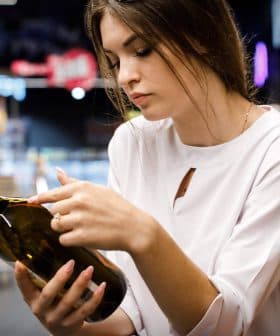 Woman inspecting a dark glass bottle of olive oil while shopping in a grocery store aisle. - Olive Oil Times