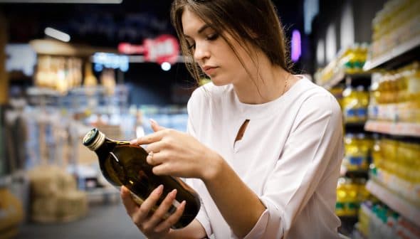 Woman inspecting a dark glass bottle of olive oil while shopping in a grocery store aisle. - Olive Oil Times