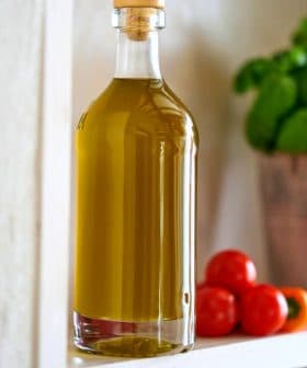 Glass bottle filled with olive oil placed on a shelf next to cherry tomatoes and a basil plant. - Olive Oil Times