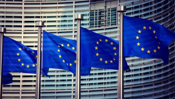 Four European Union flags displayed on poles in front of a modern building. - Olive Oil Times