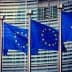 Four European Union flags displayed on poles in front of a modern building. - Olive Oil Times