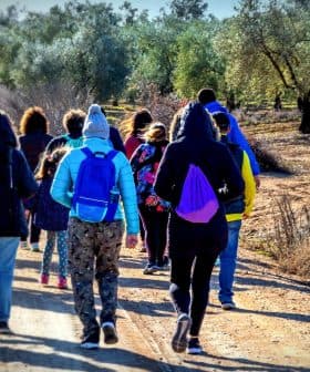 A group of people walking along a dirt path in an olive grove, wearing winter clothing and backpacks. - Olive Oil Times