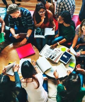 A diverse group of individuals gathered around a table, engaged in discussion with notebooks and materials. - Olive Oil Times