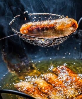 A metal strainer lifting a fried sausage from a pan of hot oil with bubbling surface. - Olive Oil Times