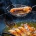A metal strainer lifting a fried sausage from a pan of hot oil with bubbling surface. - Olive Oil Times