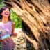 Woman with dark hair laughing while holding a bottle near an olive tree. - Olive Oil Times