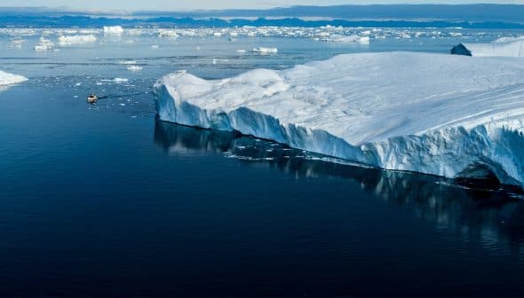 A large glacier edge with icebergs floating in calm water under a clear sky. - Olive Oil Times