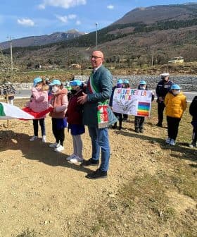 Group of children wearing blue caps at an olive tree planting ceremony with an Italian flag. - Olive Oil Times