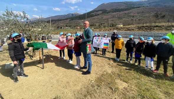 Group of children wearing blue caps at an olive tree planting ceremony with an Italian flag. - Olive Oil Times