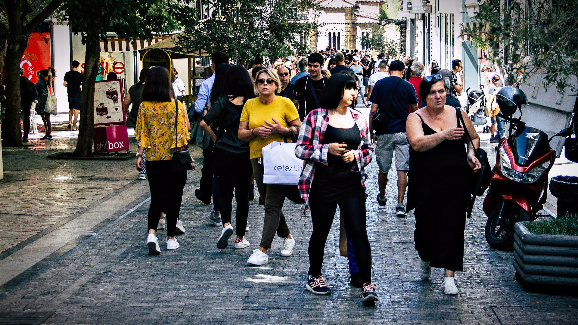 Group of people walking on a cobblestone street in an urban area with shops and trees. - Olive Oil Times