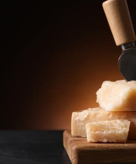Block of Parmesan cheese with a cheese knife inserted into it, resting on a wooden cutting board. - Olive Oil Times
