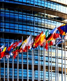 Multiple flags of European countries displayed in front of a modern glass building. - Olive Oil Times