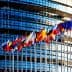 Multiple flags of European countries displayed in front of a modern glass building. - Olive Oil Times