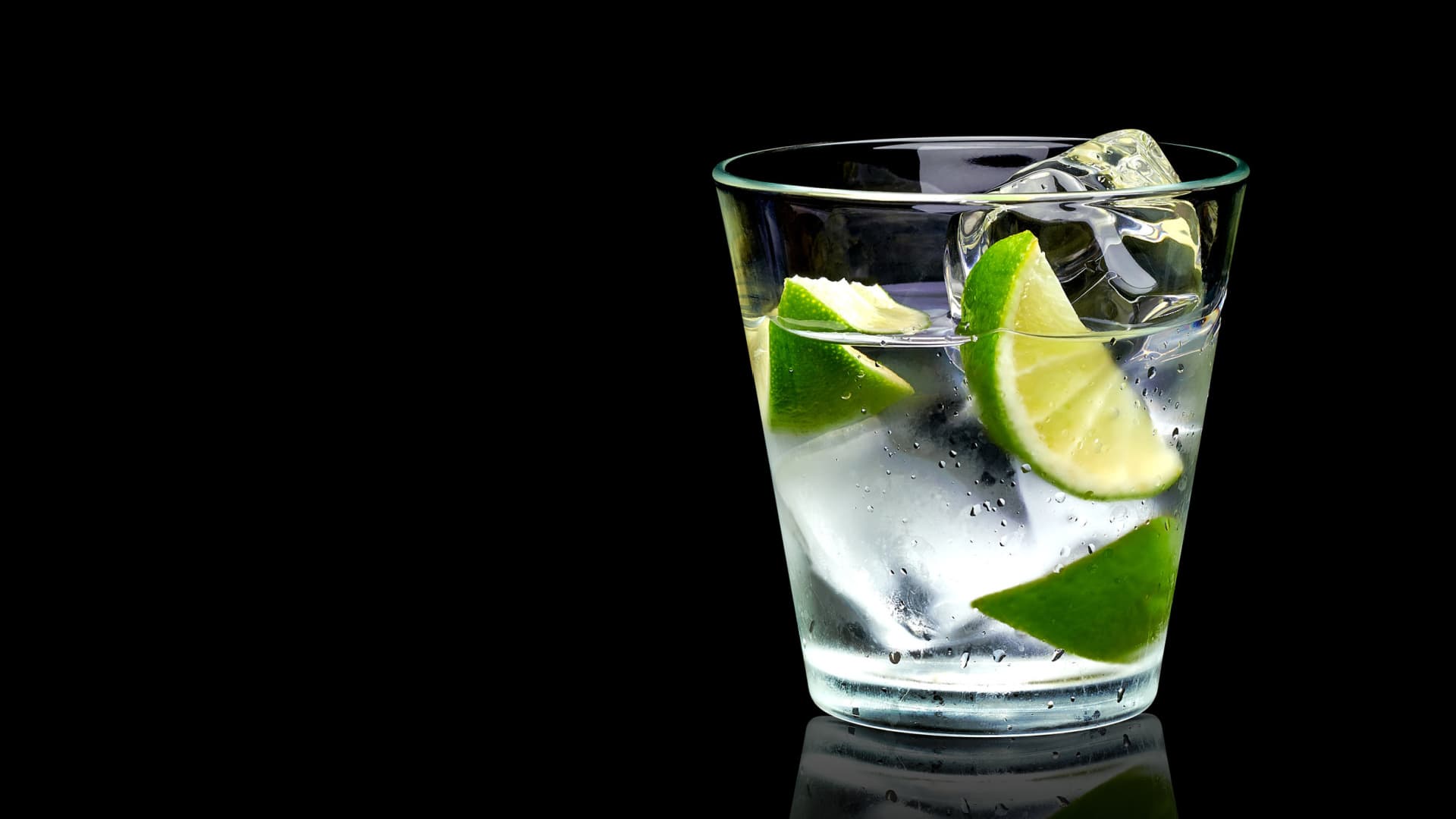 A glass filled with water, ice cubes, and lime wedges against a black background. - Olive Oil Times