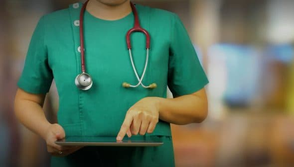 Healthcare professional in green scrubs using a tablet device while wearing a stethoscope. - Olive Oil Times