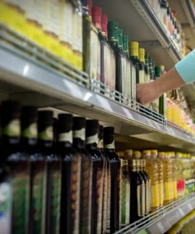 A person reaching for a bottle of olive oil on a grocery store shelf filled with various oils. - Olive Oil Times