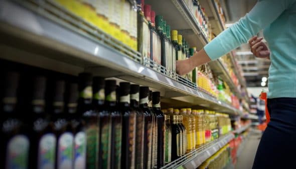A person reaching for a bottle of olive oil on a grocery store shelf filled with various oils. - Olive Oil Times