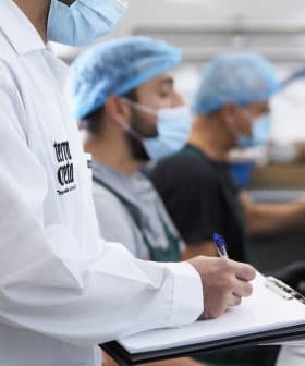 Three workers in a food processing facility wearing masks and hairnets, with one taking notes on a clipboard. - Olive Oil Times