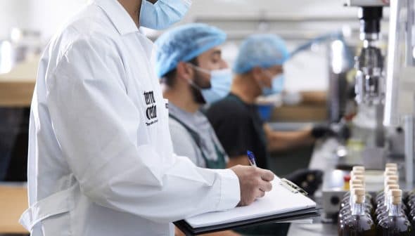 Three workers in a food processing facility wearing masks and hairnets, with one taking notes on a clipboard. - Olive Oil Times