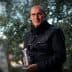 Man in a black jacket holding a bottle while standing among olive trees. - Olive Oil Times