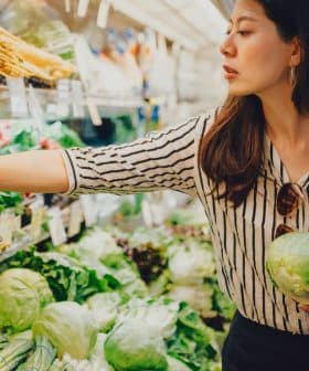 Woman selecting green onions while holding a cabbage in a grocery store produce section. - Olive Oil Times