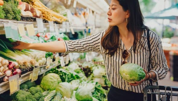 Woman selecting green onions while holding a cabbage in a grocery store produce section. - Olive Oil Times