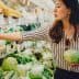 Woman selecting green onions while holding a cabbage in a grocery store produce section. - Olive Oil Times