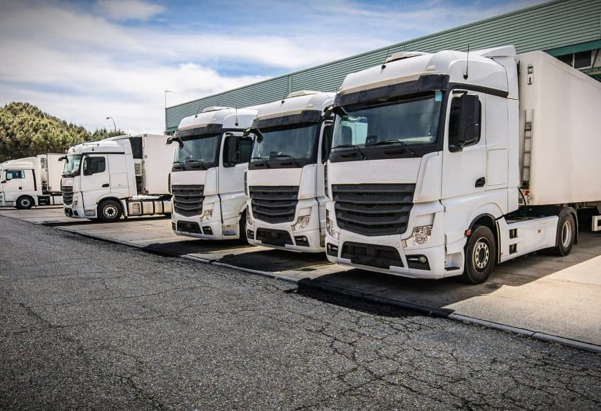 Four white delivery trucks parked in a row at a loading dock area. - Olive Oil Times