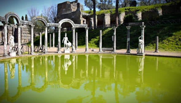 A reflective green pool surrounded by ancient Roman columns and statues in a garden setting. - Olive Oil Times
