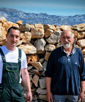 Two men posing in front of a stone wall with mountains in the background. - Olive Oil Times