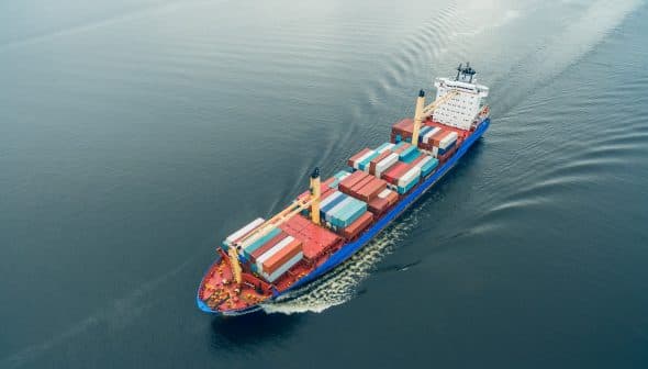 Aerial view of a container ship carrying colorful cargo containers on calm water. - Olive Oil Times