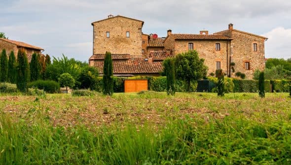 A stone building with a red roof surrounded by greenery and a field in a rural area. - Olive Oil Times