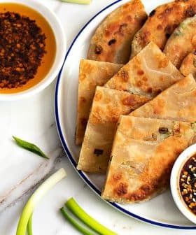 A plate of scallion pancakes arranged with two bowls of dipping sauces and green onions on a marble surface. - Olive Oil Times