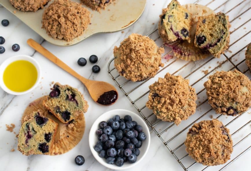 Freshly baked blueberry muffins with a crumb topping on a cooling rack and a plate of blueberries. - Olive Oil Times