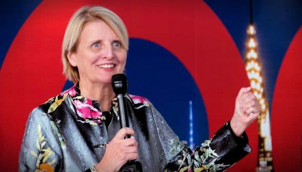 A woman with short blonde hair speaking into a microphone at an event, wearing a floral-patterned jacket. - Olive Oil Times