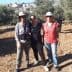 Three individuals standing together in an olive grove, wearing hats and casual clothing. - Olive Oil Times
