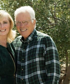 A couple smiling while posing outdoors in front of olive trees, wearing casual clothing. - Olive Oil Times