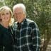 A couple smiling while posing outdoors in front of olive trees, wearing casual clothing. - Olive Oil Times