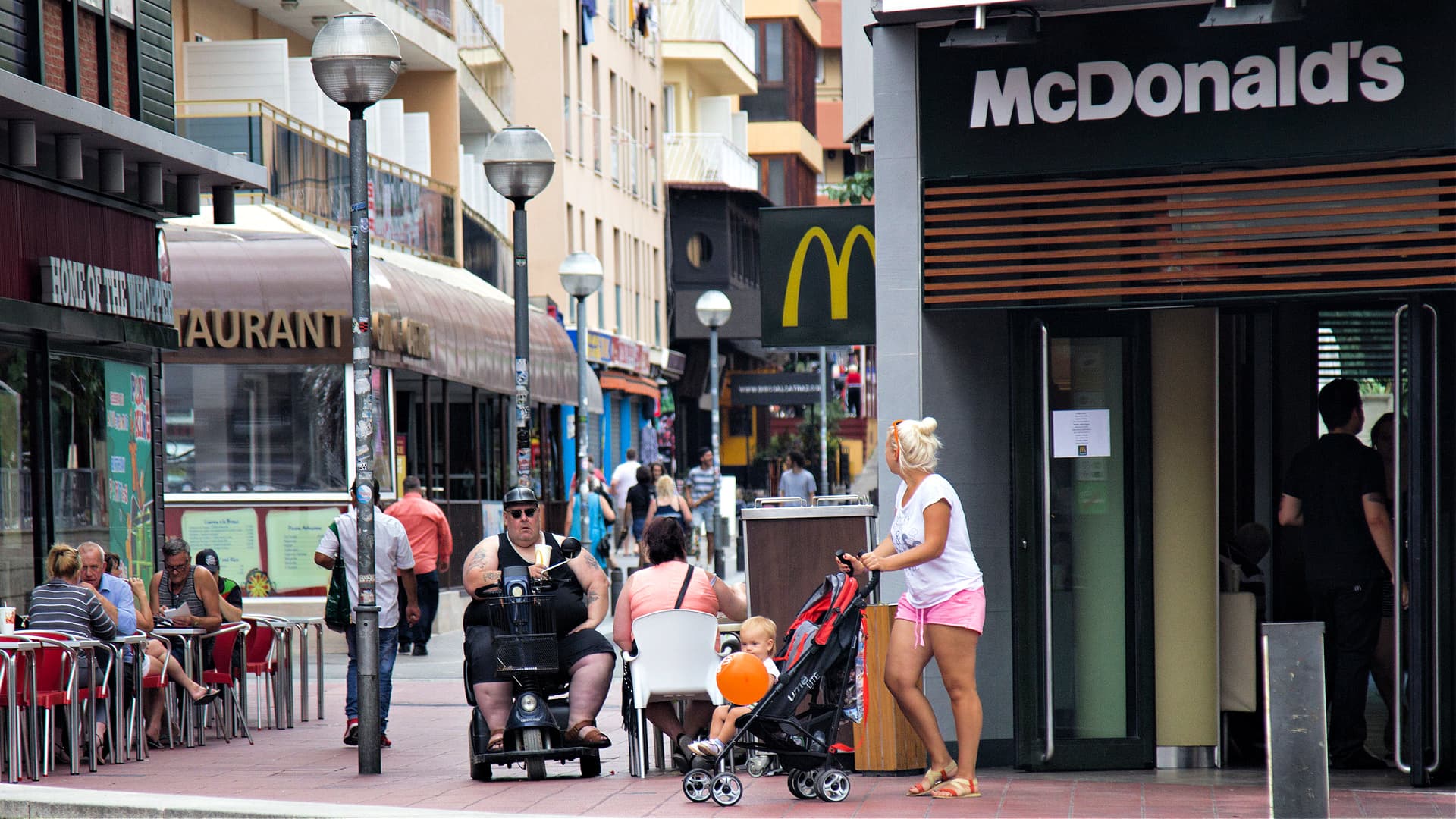 A street scene featuring a McDonald's restaurant and pedestrians, including a woman with a stroller and a man on a mobility scooter. - Olive Oil Times