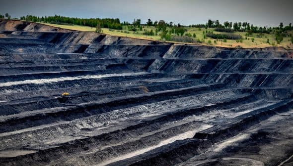 Aerial view of an open-pit mining site with layered black earth and machinery in the distance. - Olive Oil Times