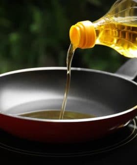 A bottle of cooking oil being poured into a red frying pan on a stovetop. - Olive Oil Times
