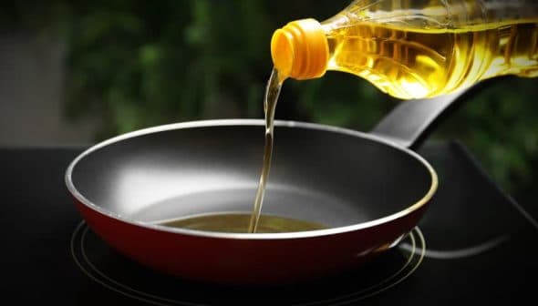 A bottle of cooking oil being poured into a red frying pan on a stovetop. - Olive Oil Times