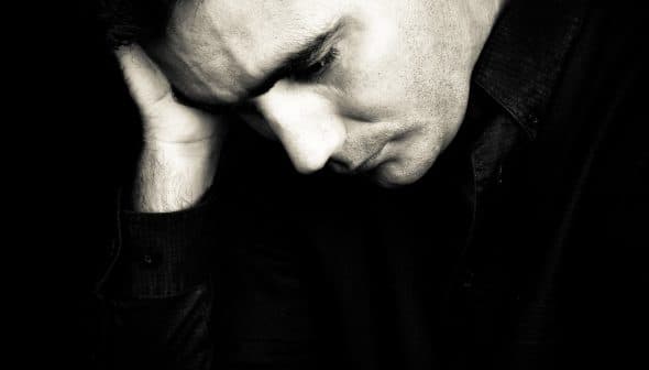 A man in a black shirt resting his head on his hand, appearing contemplative in black and white. - Olive Oil Times