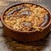 A baked dish containing sausages, onions, and grains in a round ceramic container on a wooden surface. - Olive Oil Times