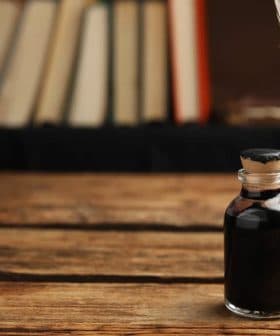A glass ink bottle with a cork stopper and a quill pen resting in a holder on a wooden table. - Olive Oil Times
